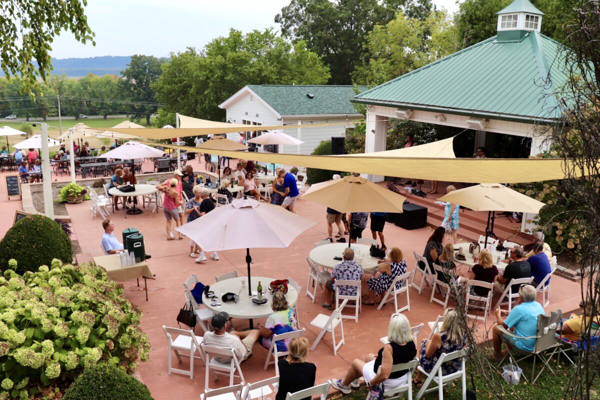 Aerial view of people hanging out and dancing on the patio outside the winery. Green hills and a blue sky line the background.
