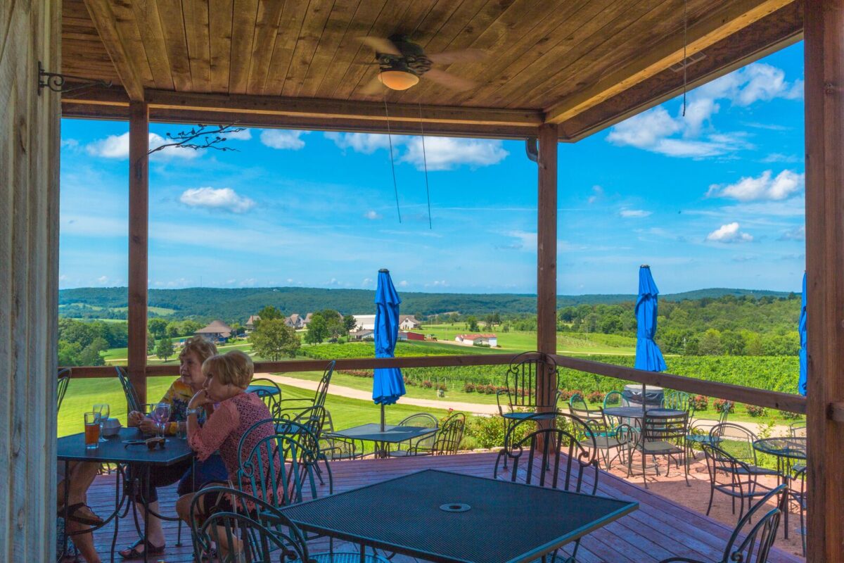 Women sit at a table on the deck while drinking wine and beer. In the background are rolling hills covered with green trees and the sky is bright blue with wispy clouds scattered throughout.