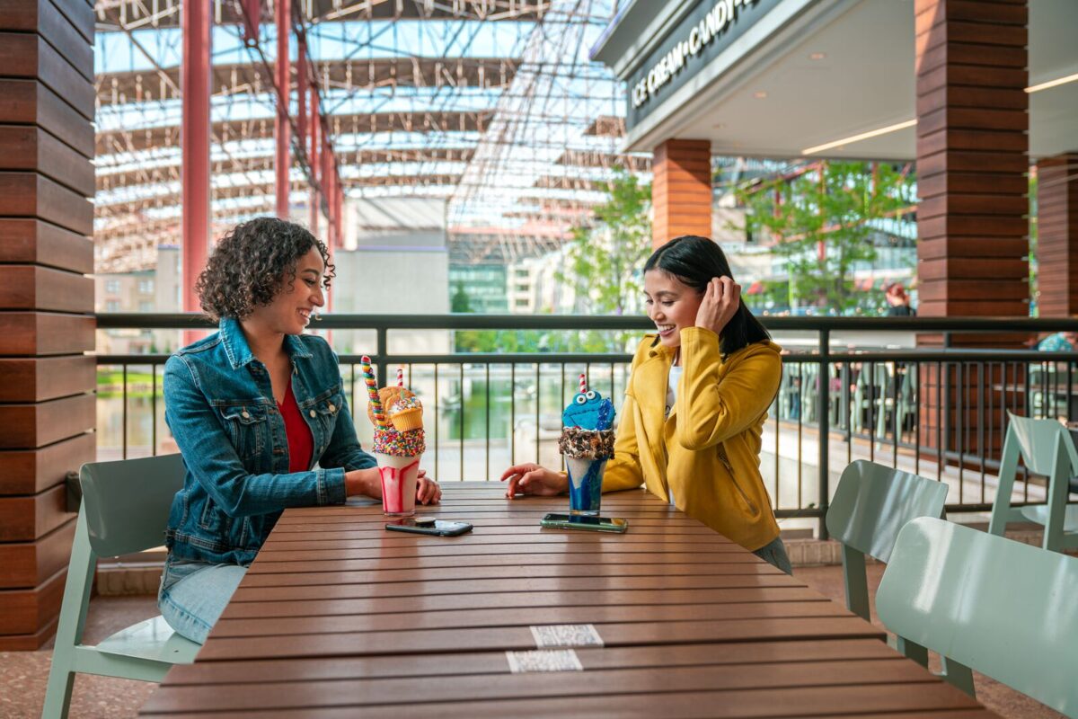 Two girls sit at a table smiling and laughing. They have fun, creatively designed milkshakes they're about to enjoy.
