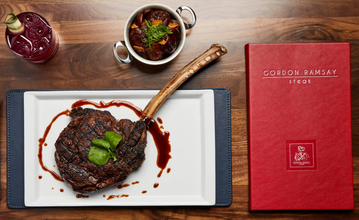 A gourmet tomahawk ribeye steak plated with sauce and garnish, served alongside a cocktail and side dish, with a red Gordan Ramsey Steak menu on the table.