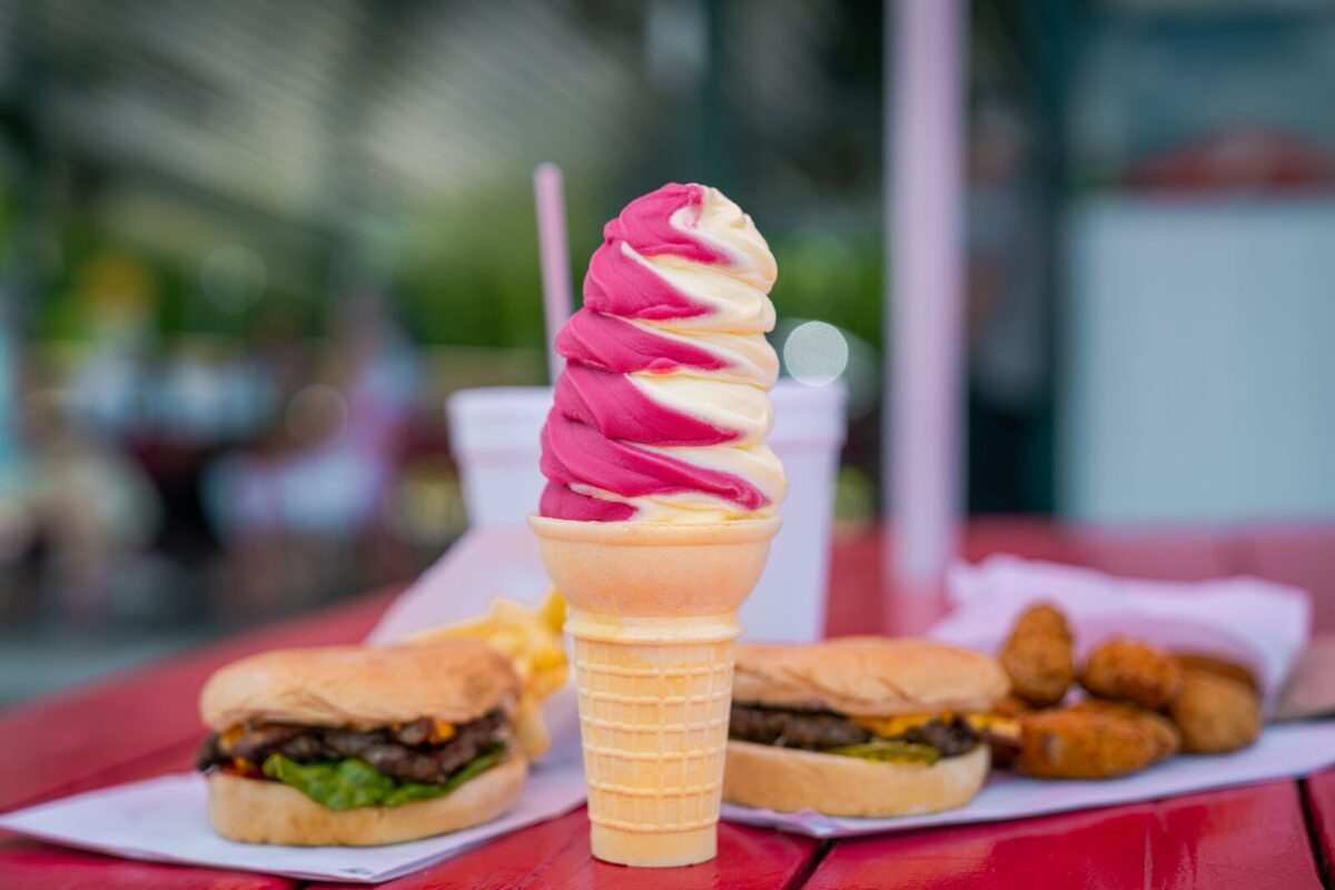 A pink and white swirl of soft-serve ice cream sits on a cake cone with burgers and drinks in the background.
