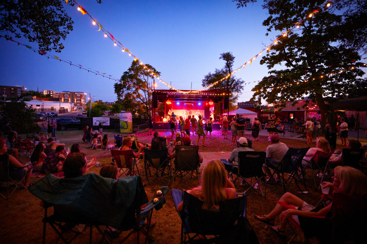 People sit in chairs outside as the glow from stage lights illuminates a crowd dancing to a live band.