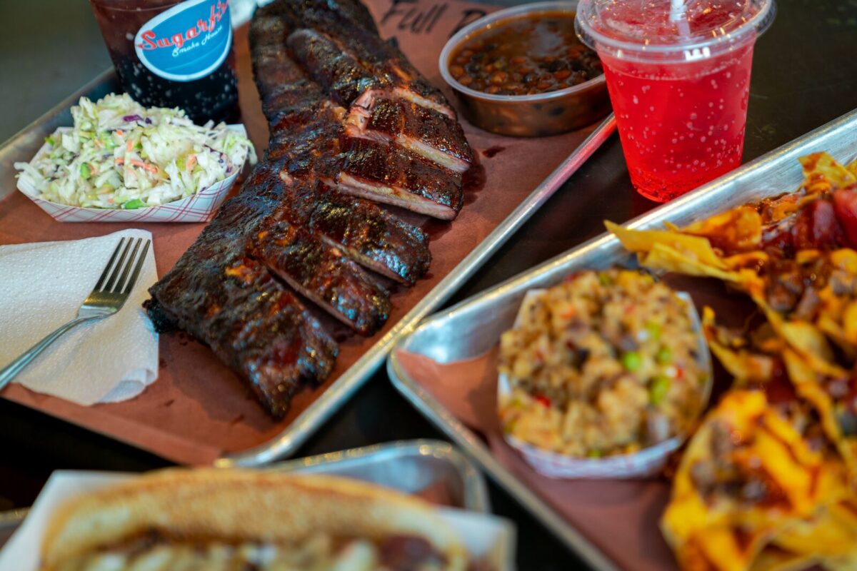 A tray full of food from Sugarfire Smokehouse features a saucy rack of ribs, a side of creamy coleslaw, loaded BBQ nachos with beef  and two cold drinks, including a bottle of root beer.