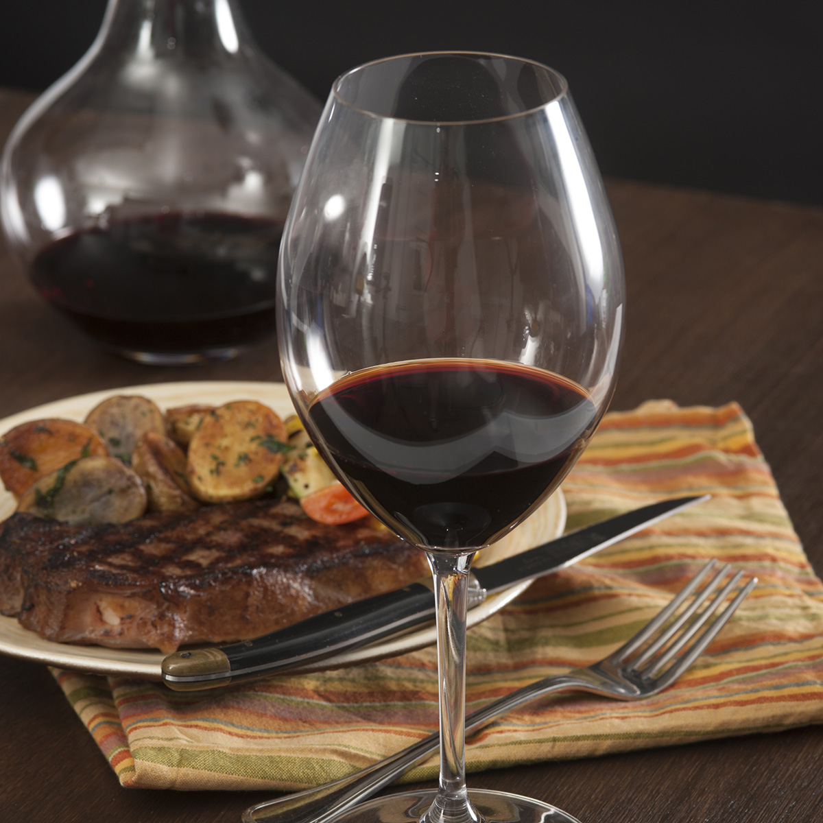 A glass of red wine on a table. In the background, a steak with potatoes and silverware and a wine carafe are partially visible.