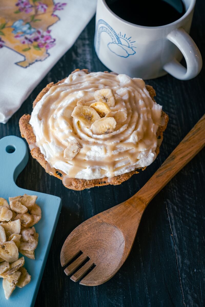 A pie topped with bananas sits on a table with a wooden spoon, napkin , mug of coffee and a cutting board with banana slices.