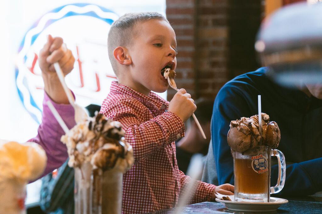 A young boy licks his spoon as he enjoys a large ice cream float. 