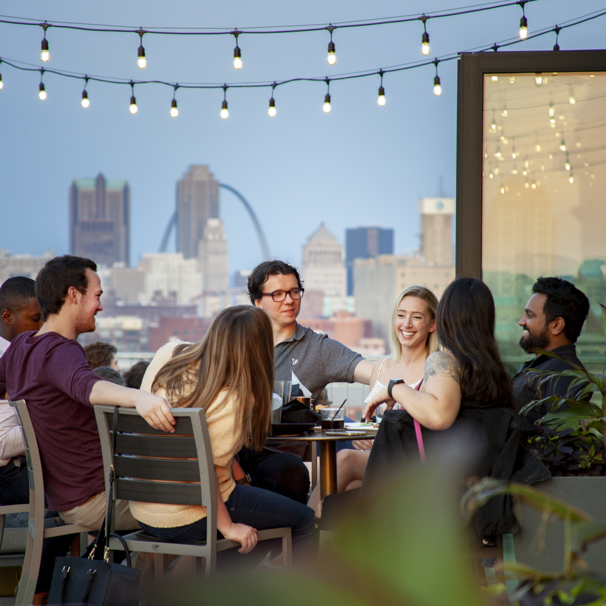 A group of people sit on a rooftop bar and enjoy drinks with the Gateway Arch and downtown St. Louis in the background. 