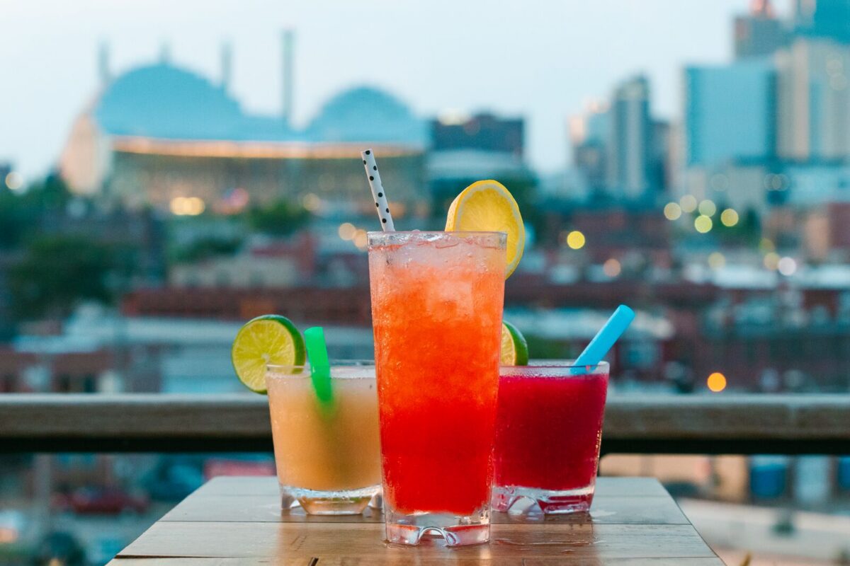 Colorful cocktails sit on a table at a rooftop bar with a city skyline in the background.