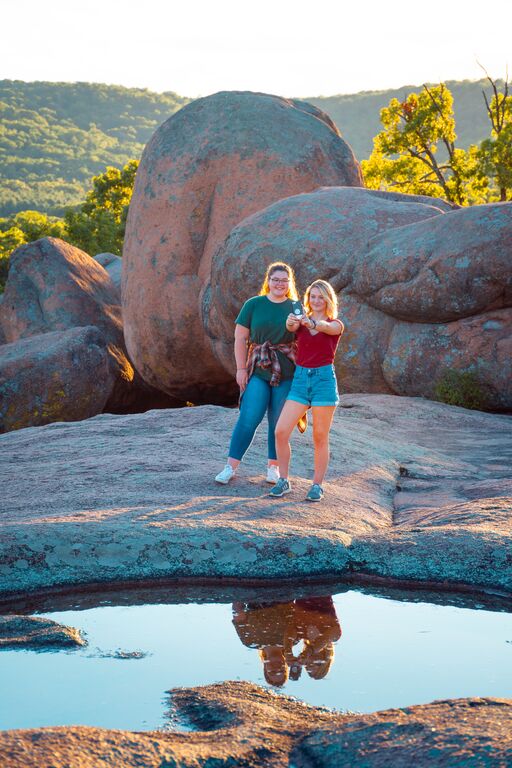 Two people pose for a selfie outside large boulders on a hiking trail.