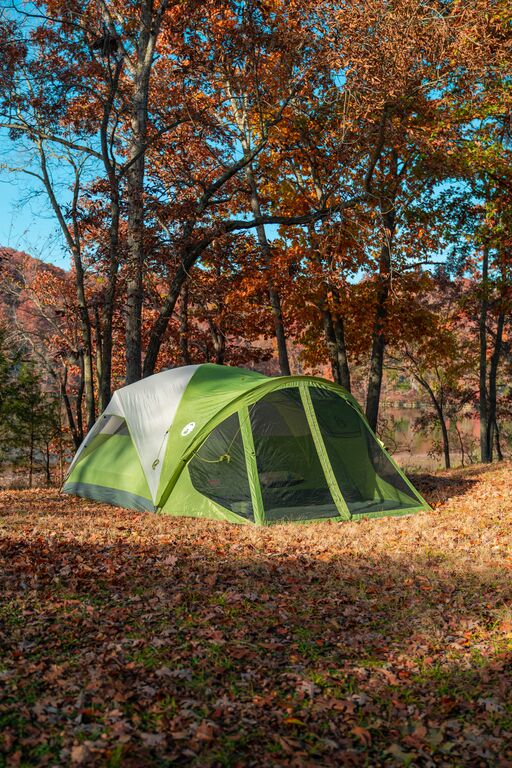 A green tent sits in a leaf covered site with fall leaves in the background.