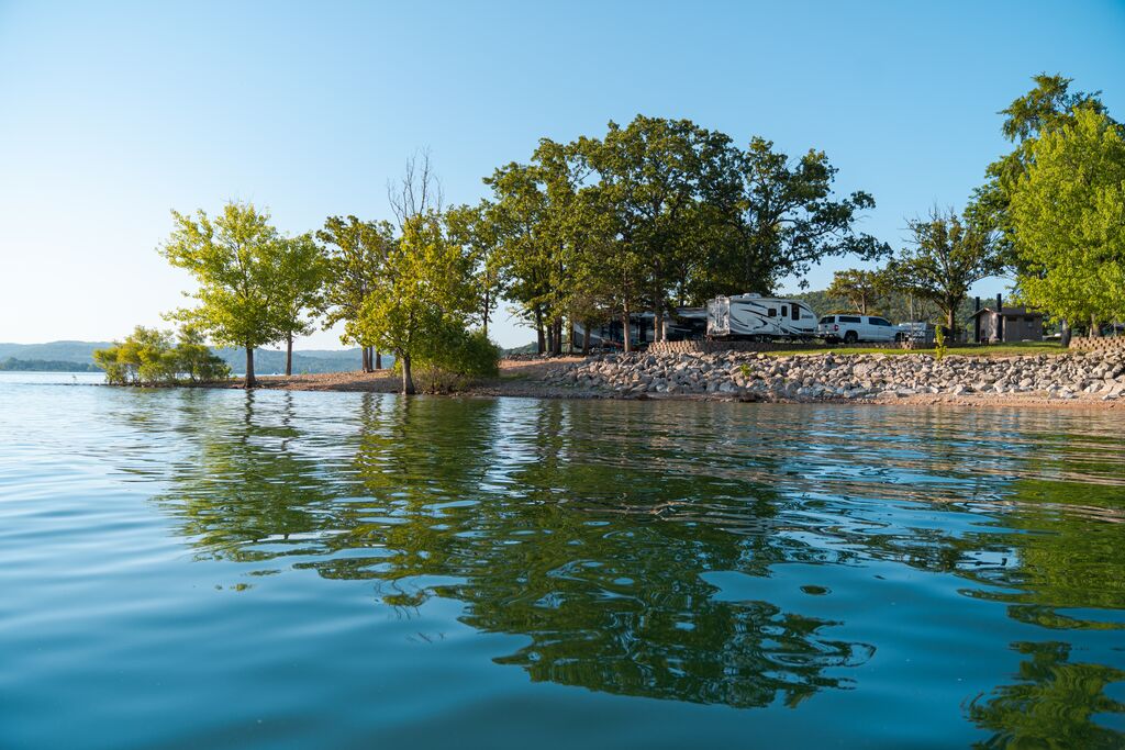 Campers, vehicles and structures on the rocky bank of a blue and green lake with a clear sky overhead. 