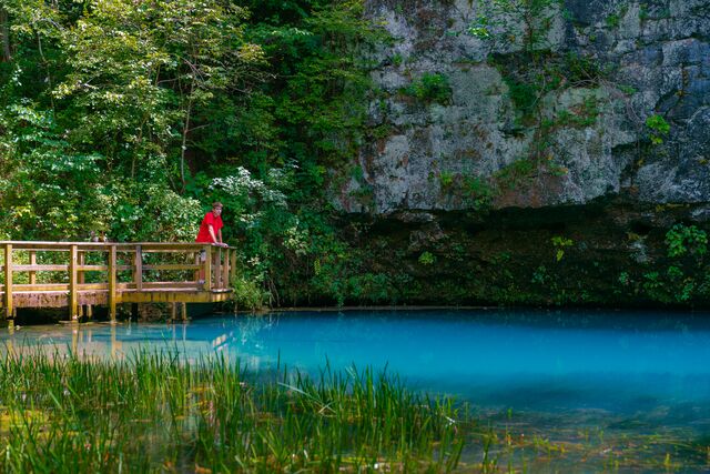 A teen boy standing on the edge of a deck looks out over Blue Spring, a vivid turquoise pool of water surrounded by rock bluffs and a lush green forest.