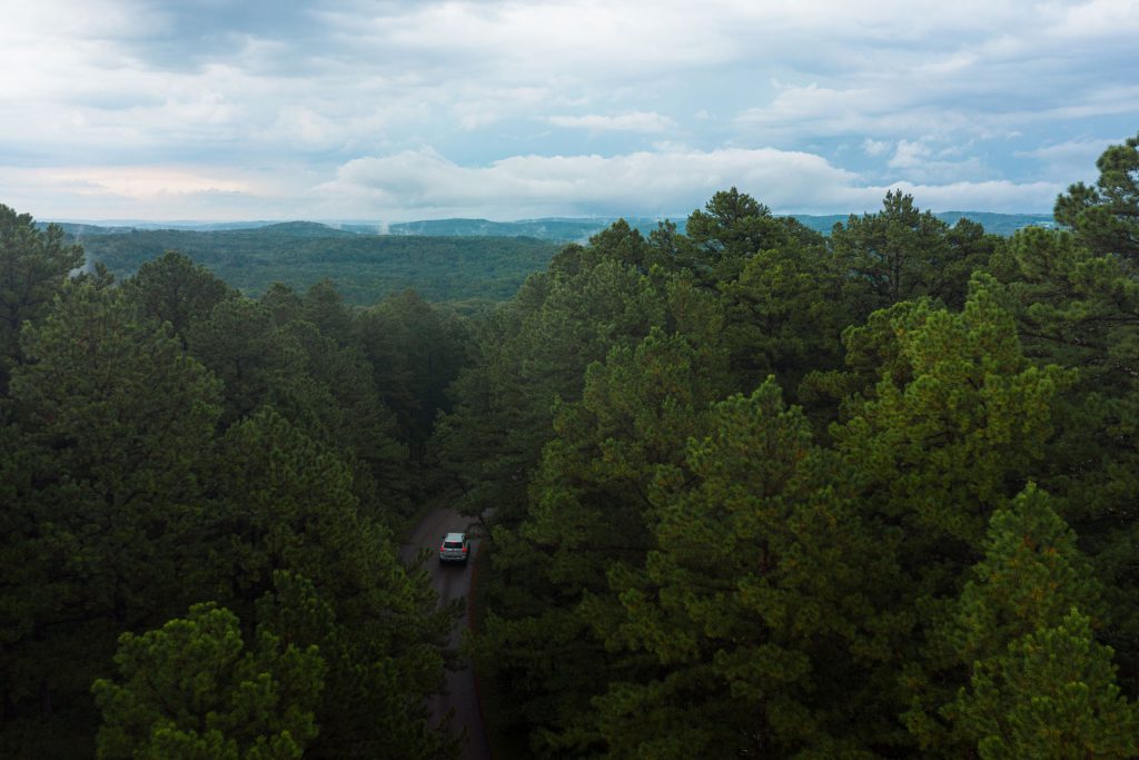 A car drives on a winding road through a forest. Rolling hills are visible in the distance.