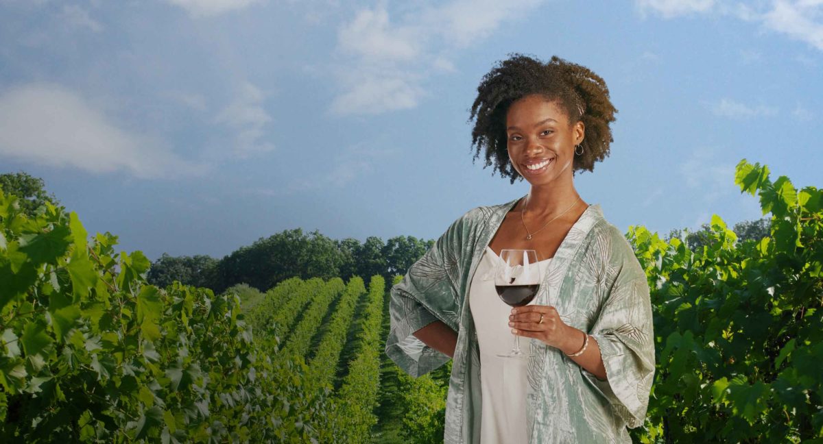 A woman stands holding a glass of red wine in a vineyard.