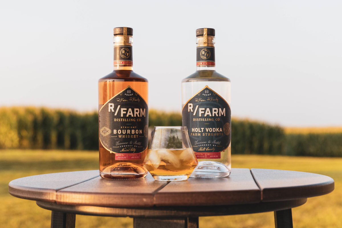 Two bottles of spirits on a table outdoors with a glass full of bourbon and ice in between them.