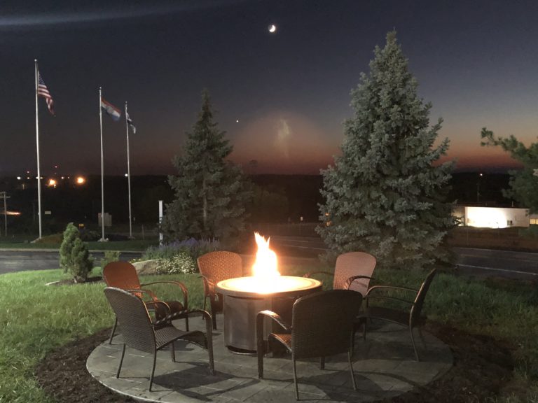 A lit fire pit with trees and chairs around it.