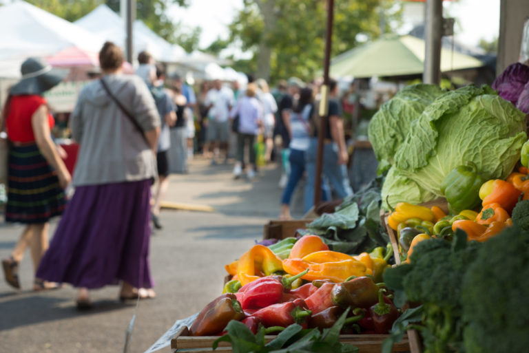 Colorful fresh produce are highlighted in the foreground with a busy farmers market filled with shoppers in the background.