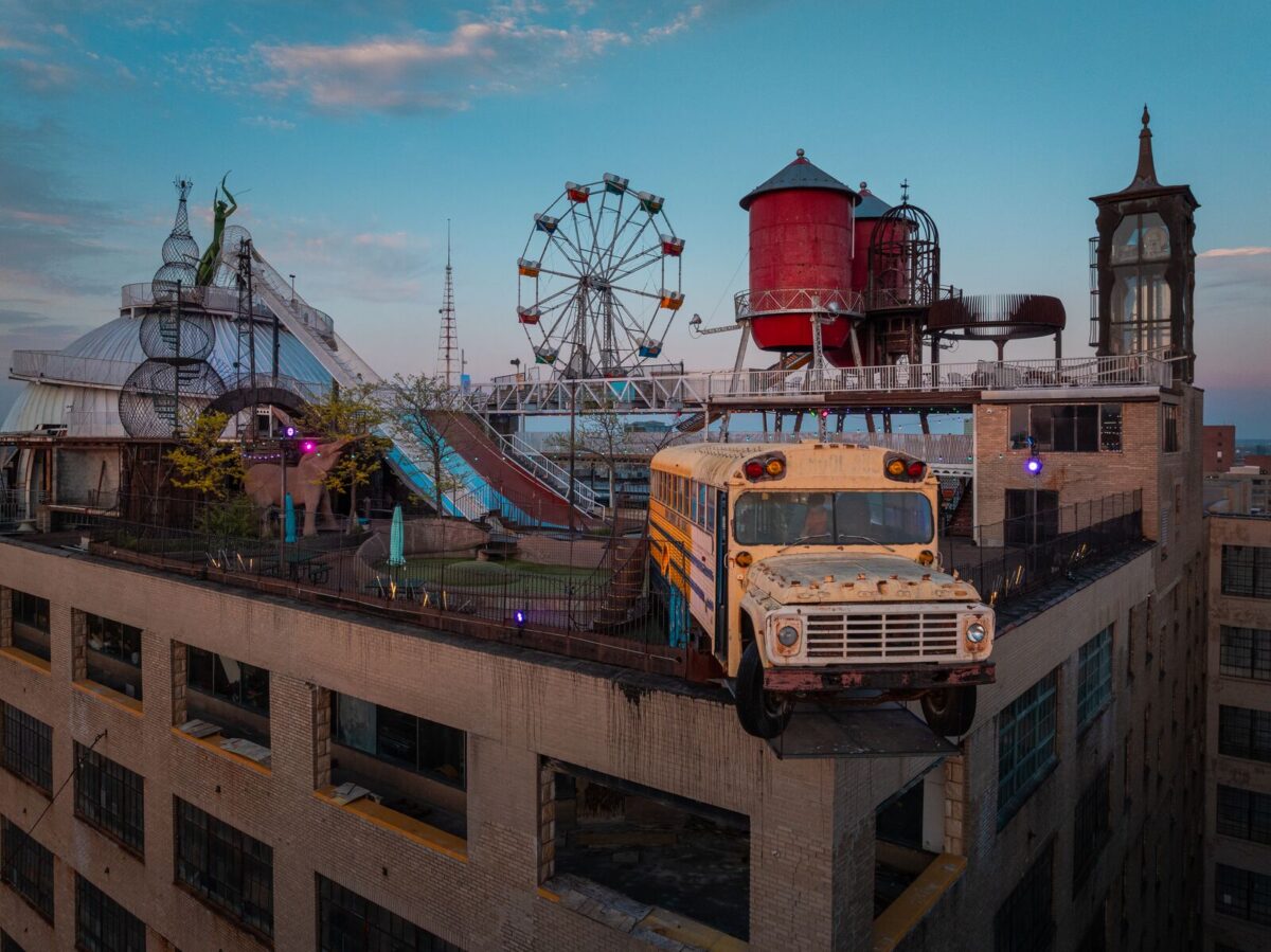 The top of a city building with a Ferris wheel, a school bus dangling from the top corner and other fun attractions.