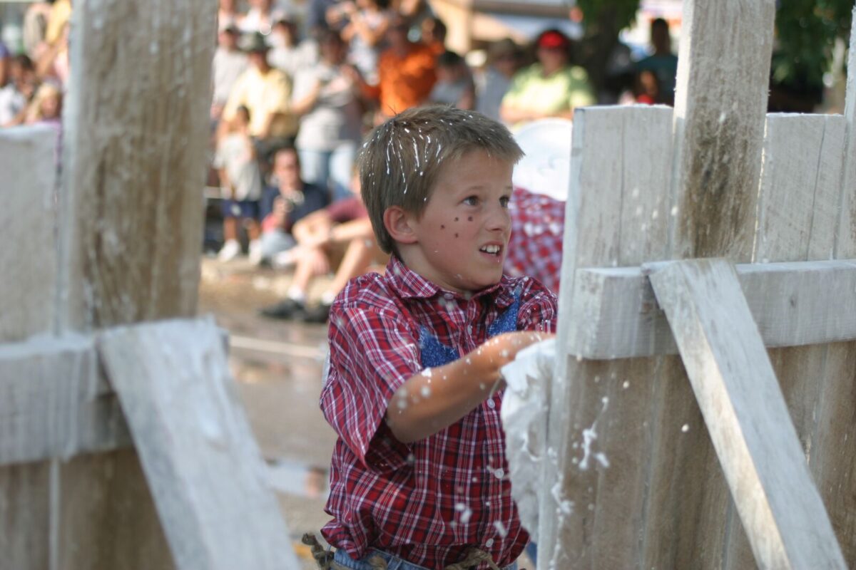A boy at a summer festival for National Tom Sawyer Days participates in a white fence painting contest. He wears a red, farm-style shirt with blue jeans.