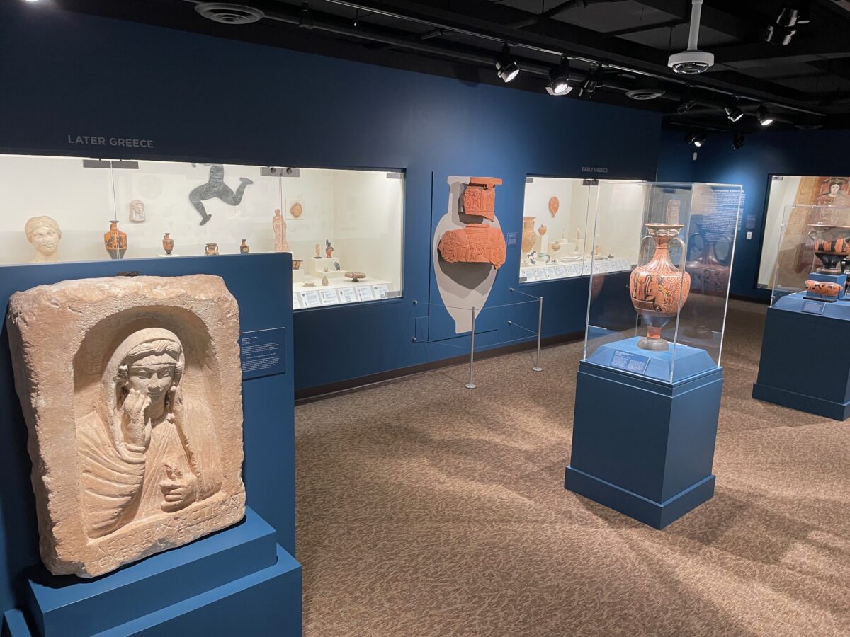 A museum with dark blue walls features artifacts in cases in the wall and in the small cases throughout the museum's floor.
