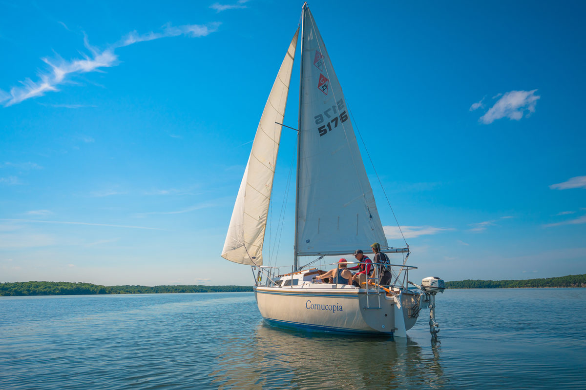 A blue, nearly cloudless sky and a calm lake with a sailboat and 3 sailors sailing through the water.