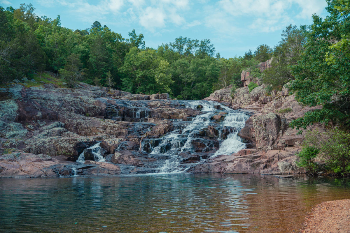 A waterfall flows over reddish-brown rock into a calm pool of water. Surrounded by lush green trees under a partly cloudy sky.