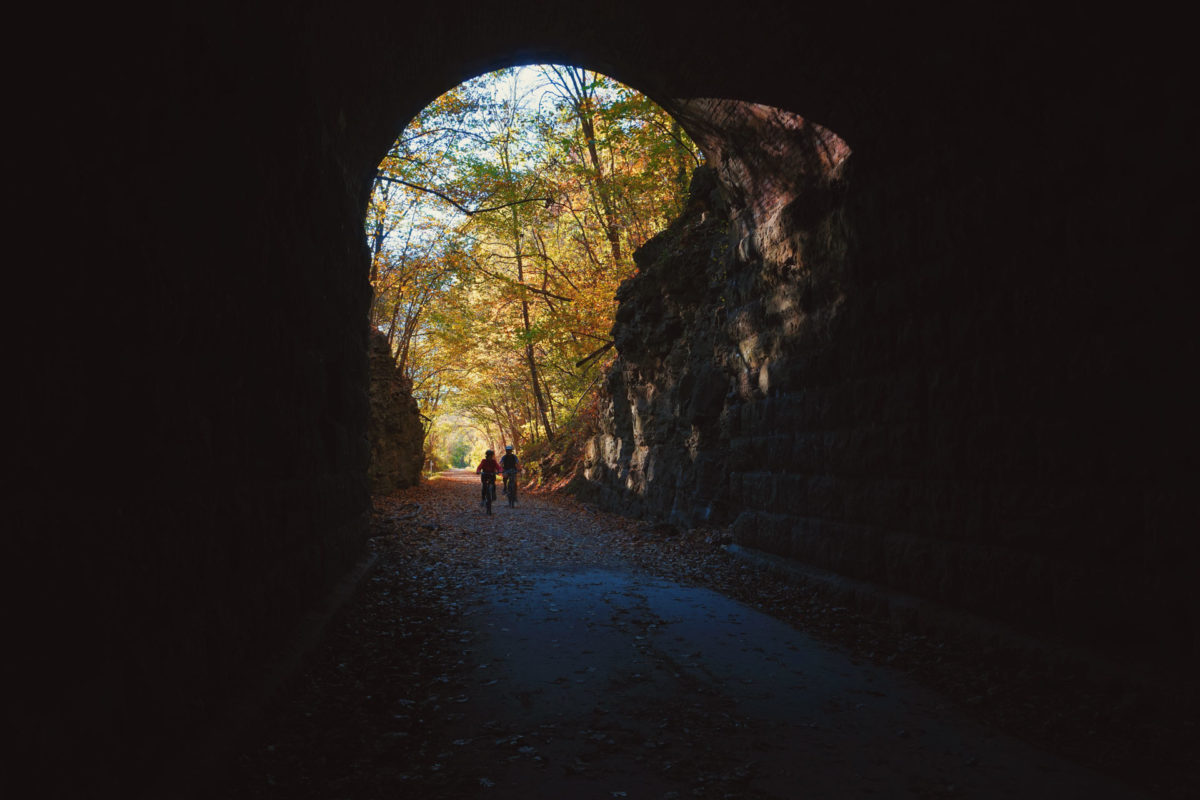 Two bicyclists wearing helmets ride from a fall leaf-covered trail into a dark tunnel.