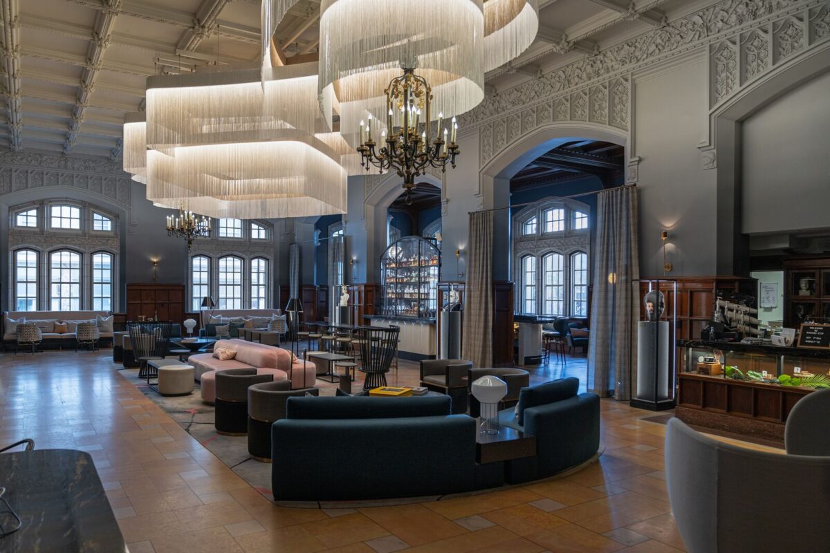 The interior of Hotel Kansas City features ornate wall and ceiling details, a large, curving white light fixture, a gold chandelier, and velvet couches.