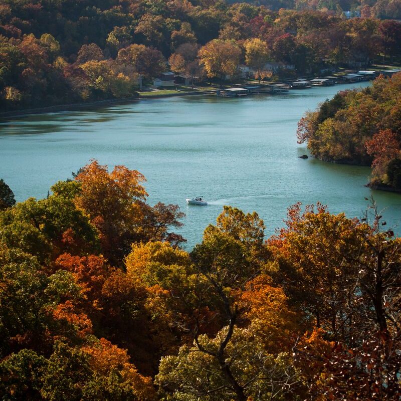 A boat drives through the Lake of the Ozarks with colorful fall foliage lining the coast.