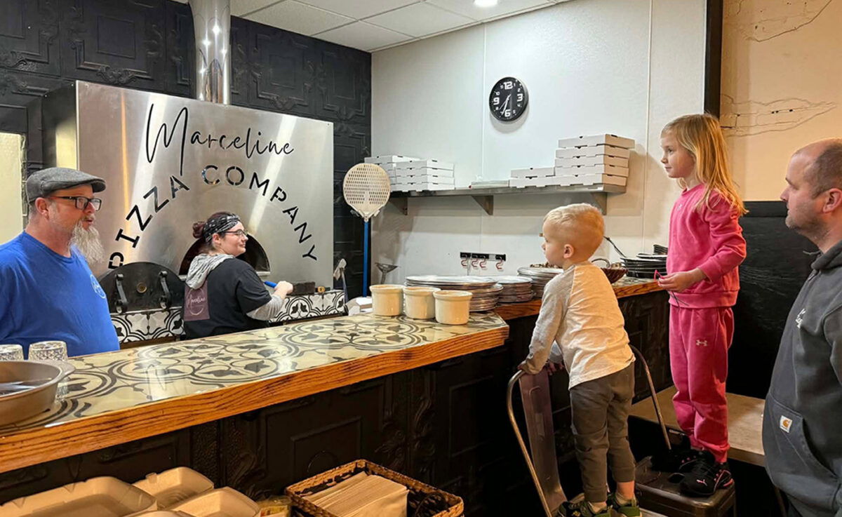 Children near the counter of a pizza restaurant with adults working on the other side.