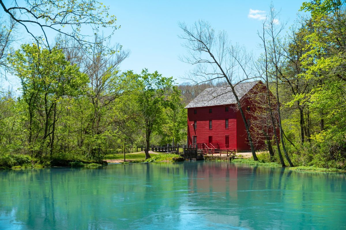 A red mill overlooks a turquoise pond with green trees all around.