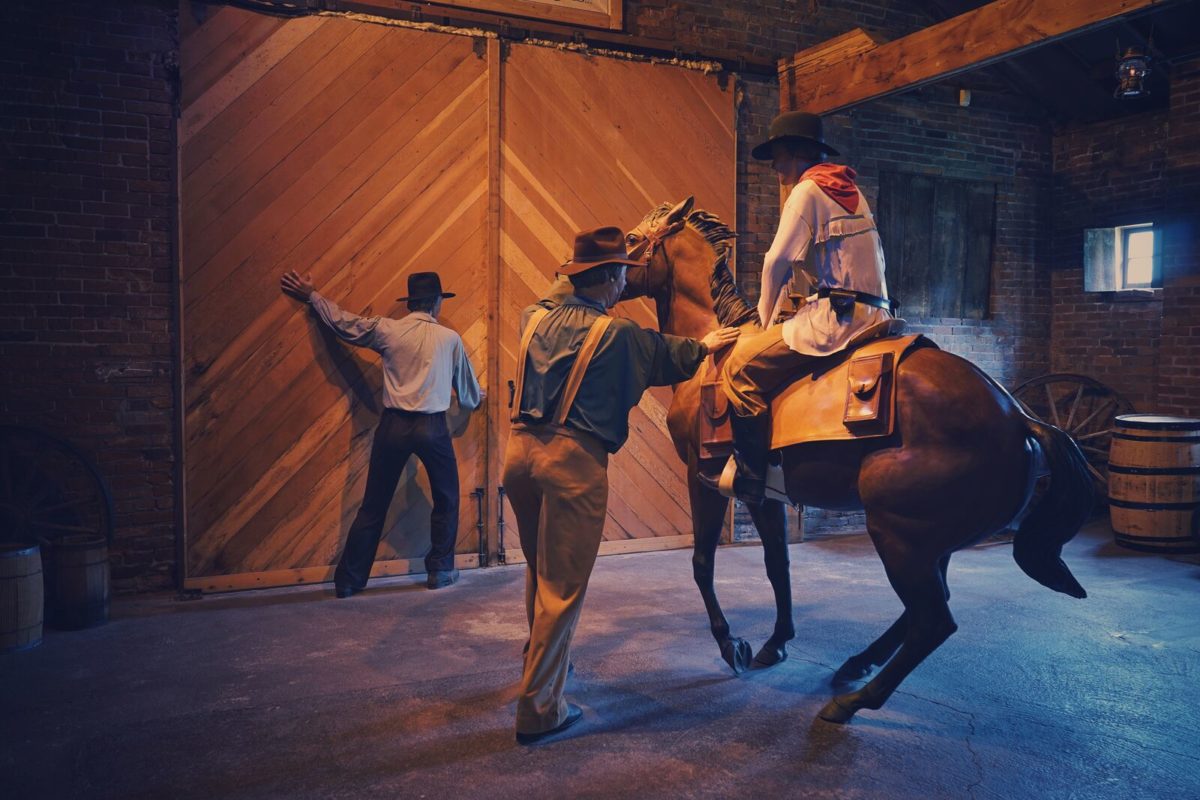 Three figures stand in a tableau — one figure is seated on a horse while a second assists. The third opens a wooden barn door.