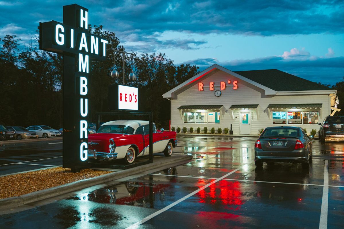 A classic car sits in a wet parking lot reflecting the neon light of multiple signs. 