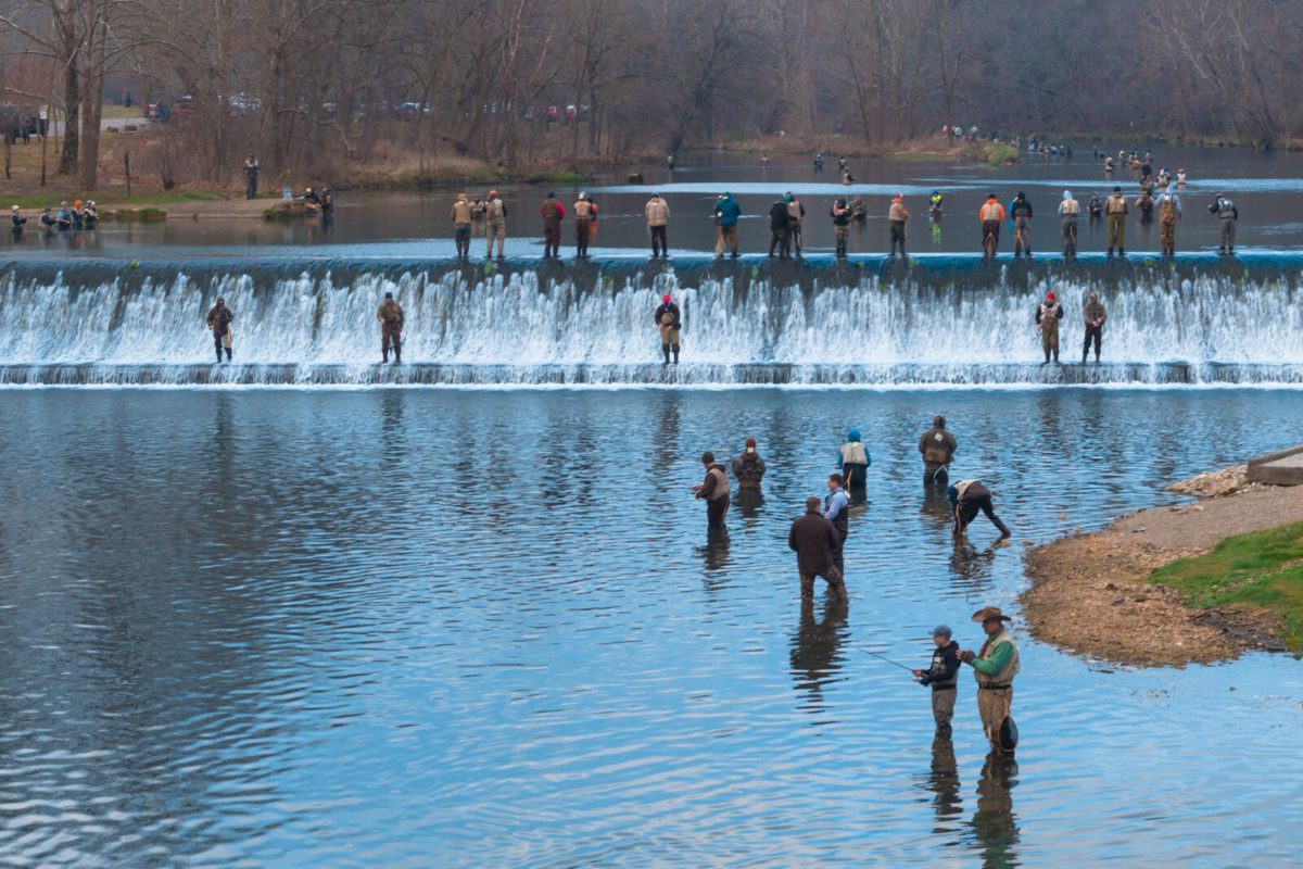 A line of people fish in a cold river with a small waterfall.