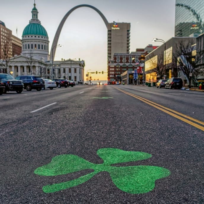 A shamrock is painted on the street with the Gateway Arch in the background. 