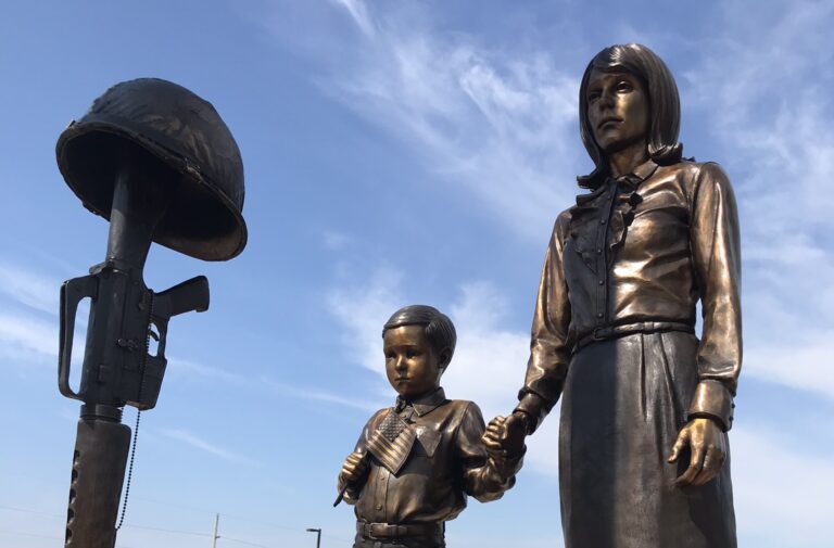 A sculpture of a woman and child, holding an American flag near their heart, gazing at a rifle turned nose-down with a helmet on its stock.