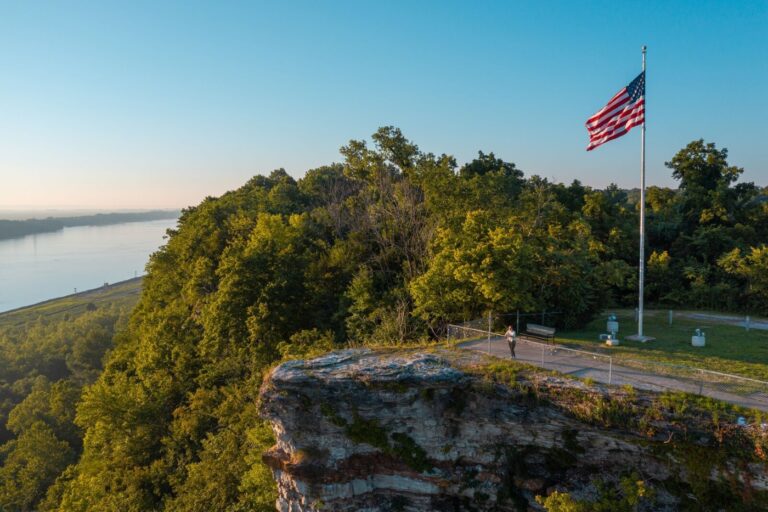 An aerial view shows a person looking out over a bluff and forested hills. The Mississippi River is in the background, and the sky is a bright blue with no clouds.