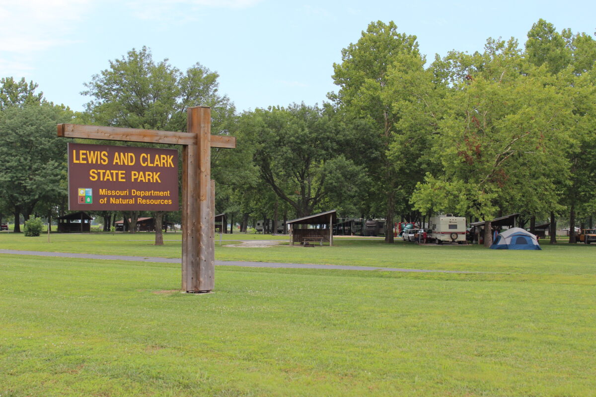 Brown and yellow Lewis and Clark State Park cantilever sign with RVs, tent, and small shelters in background