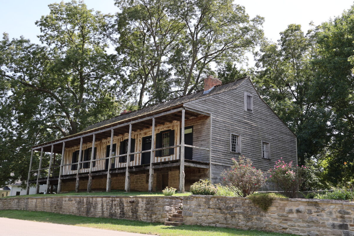 Raised home with a covered porch. Vertical log construction is visible. Stone foundation and wooden siding on side.