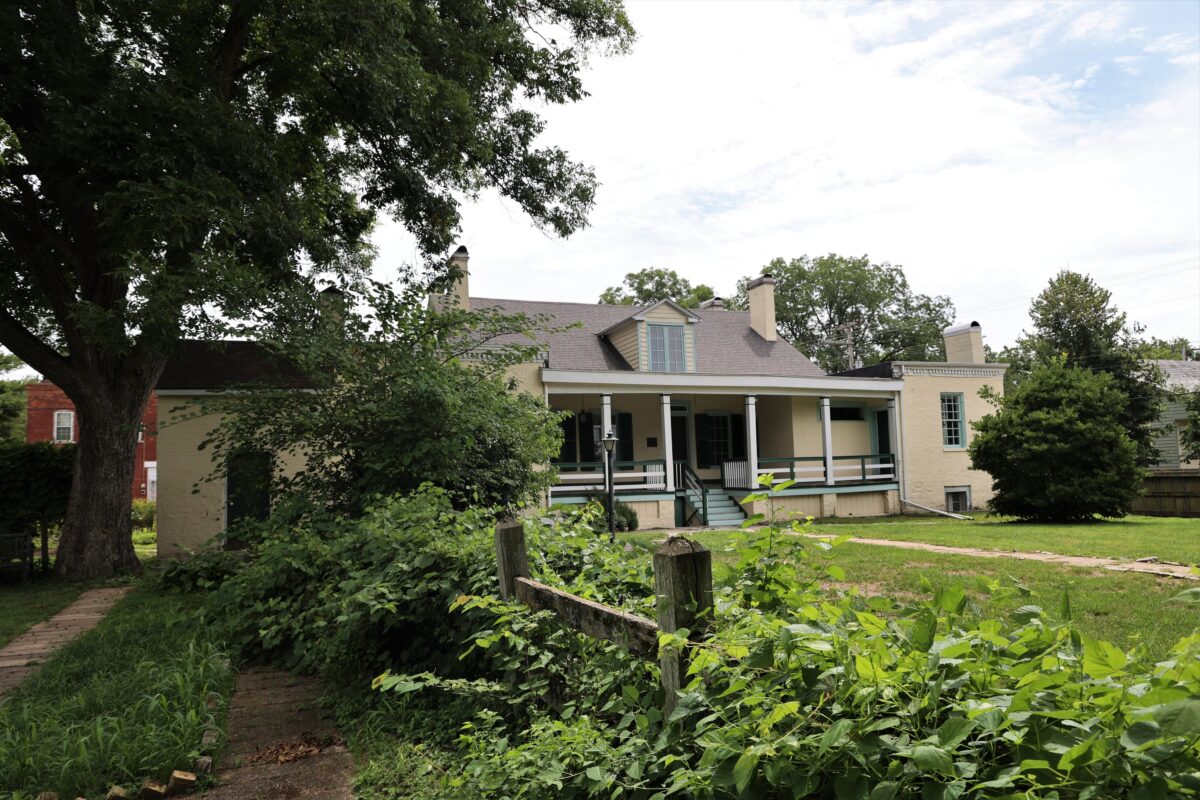 A backyard of a cream colored house with back covered porch. A fence covered in vegetation and a large tree on the left.