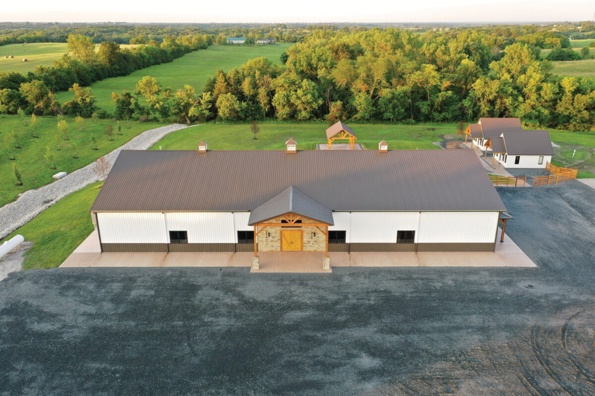 Aerial view of the front of The VeNue Event Space--a big white building with a grey roof and three cupolas. It has a grande front entrance made of stone with an overhang. Two white Villas can be viewed behind the building.