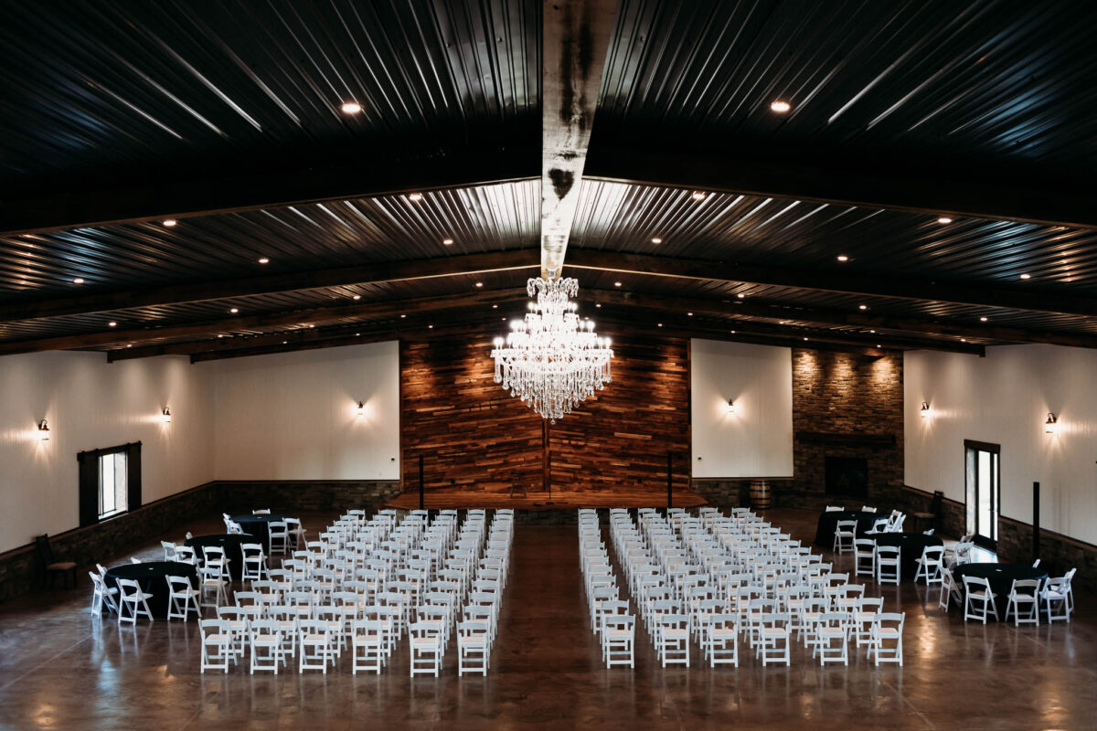 Interior photo of The VeNue Event Space set up with 300 white chairs and 6 round tables with black table clothes. A large chandelier is the focal point at the center of the main floor.