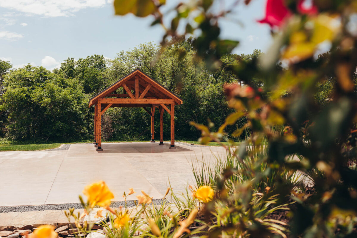A wooden pavilion on stamped concrete surrounded by a wooded area and flowered landscaping.