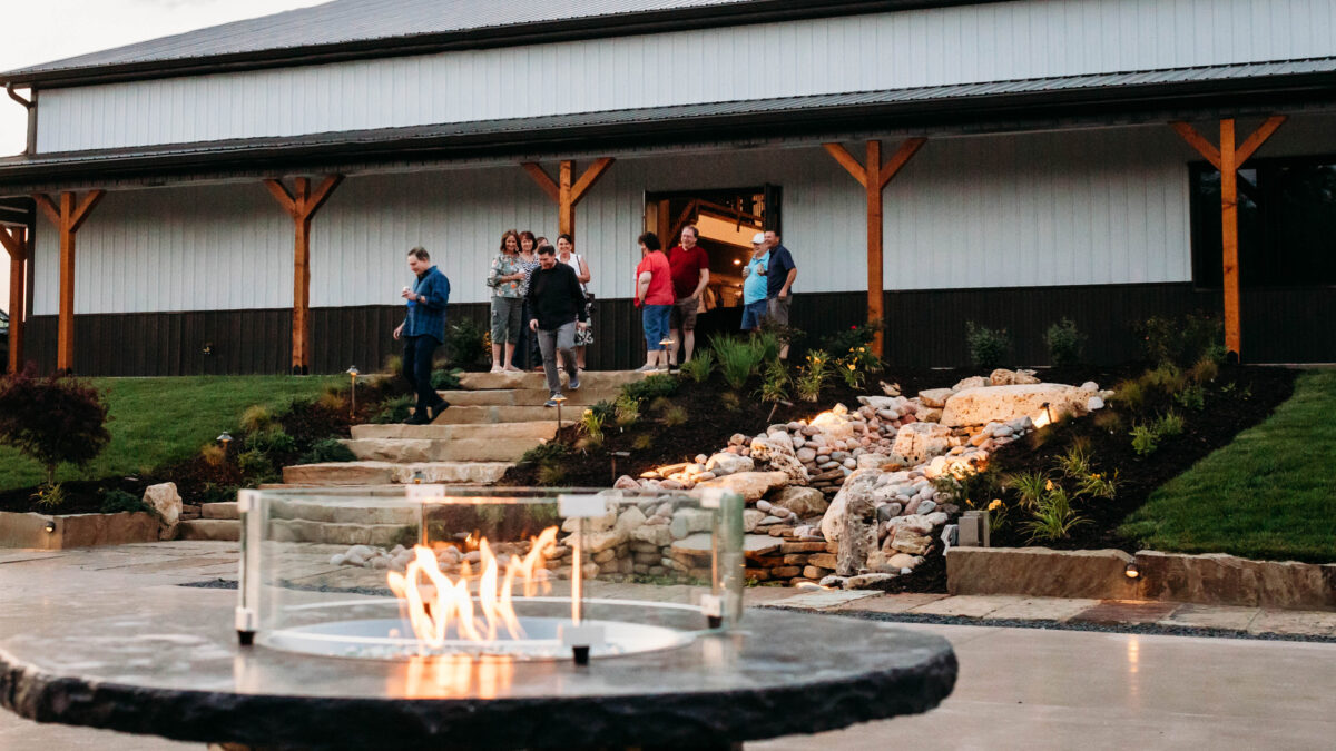Back side of The VeNue Event Space with people walking down the stone strairs. The whiskey barrel firepit is lit in the foreground.