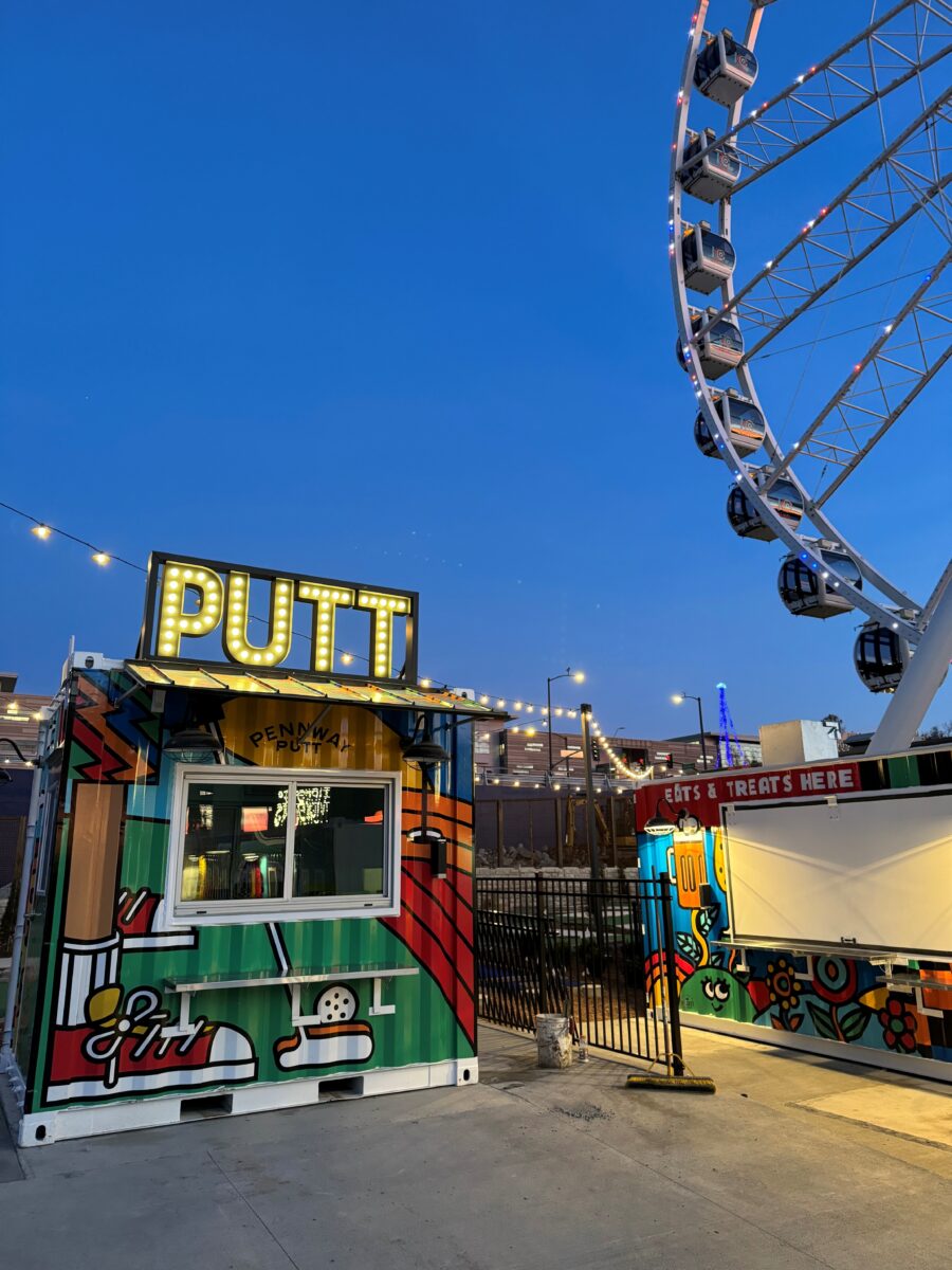 The bottom of a lit up Ferris wheel near other attractions.