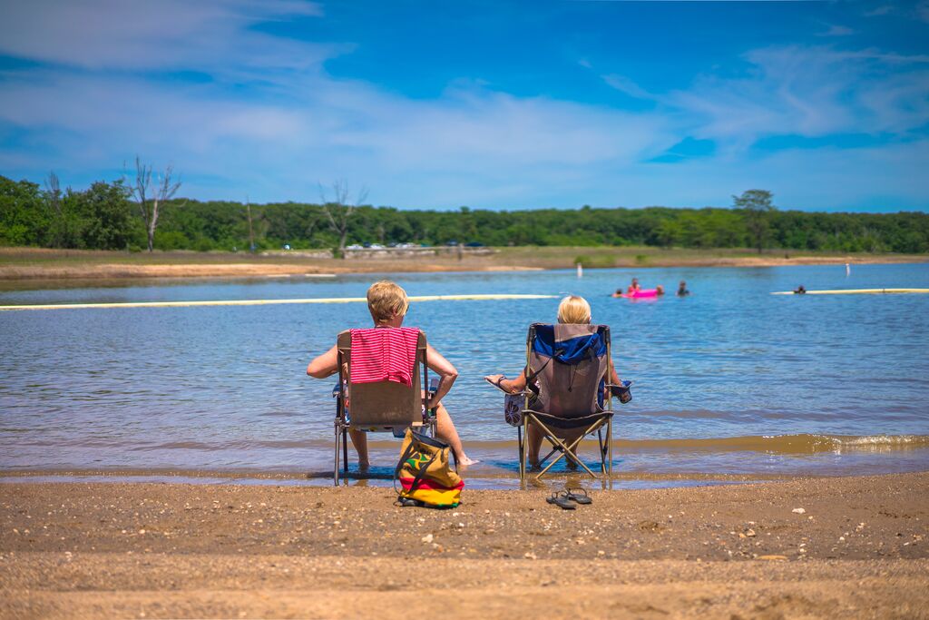 Two people sit in lawn chairs on the sand-covered bank. The tide is coming in and covering their feet. In the lake, there are children swimming and people floating. In the distance, there are lush, green trees lining the bank, and wispy clouds float throughout the blue sky.