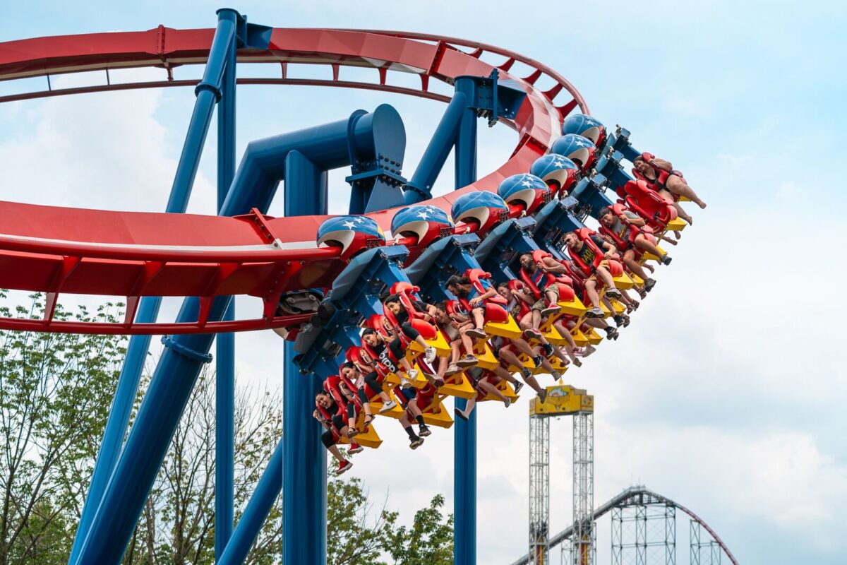 A red and blue rollercoaster at Worlds of takes riders around a curve and their feet are dangling below their seats. The blue sky and green surrounding are visible as the ride twists and turns in the air.