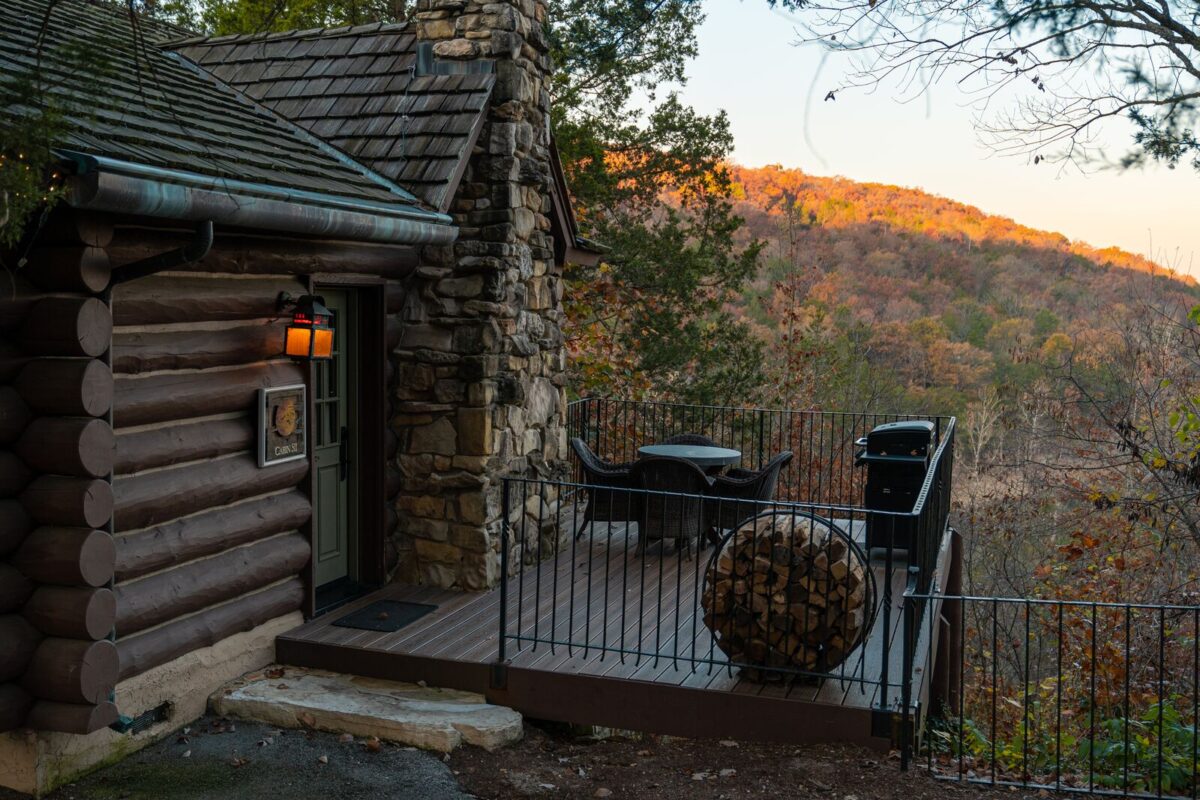 A wooden lodge in Branson sits on a hill. There is patio with seating, grill and firewood. The lodge overlooks the Ozark mountains that are shades of orange, green and yellow.