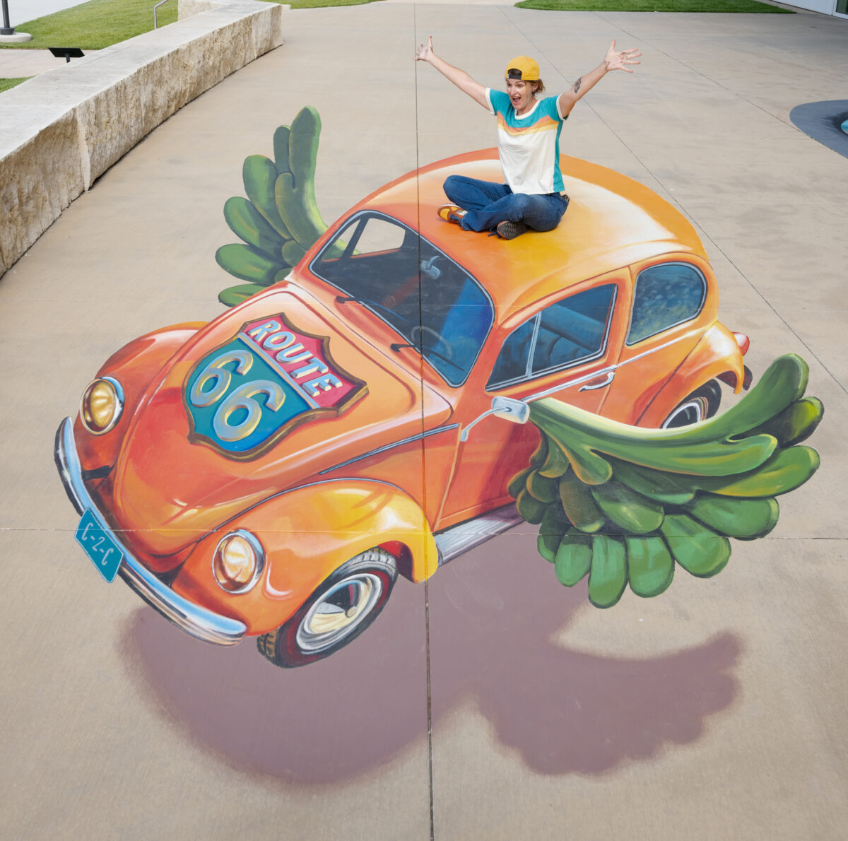 A person playfully sits on top of 3D artwork of an orange VW Beetle Route 66 car with wings. 