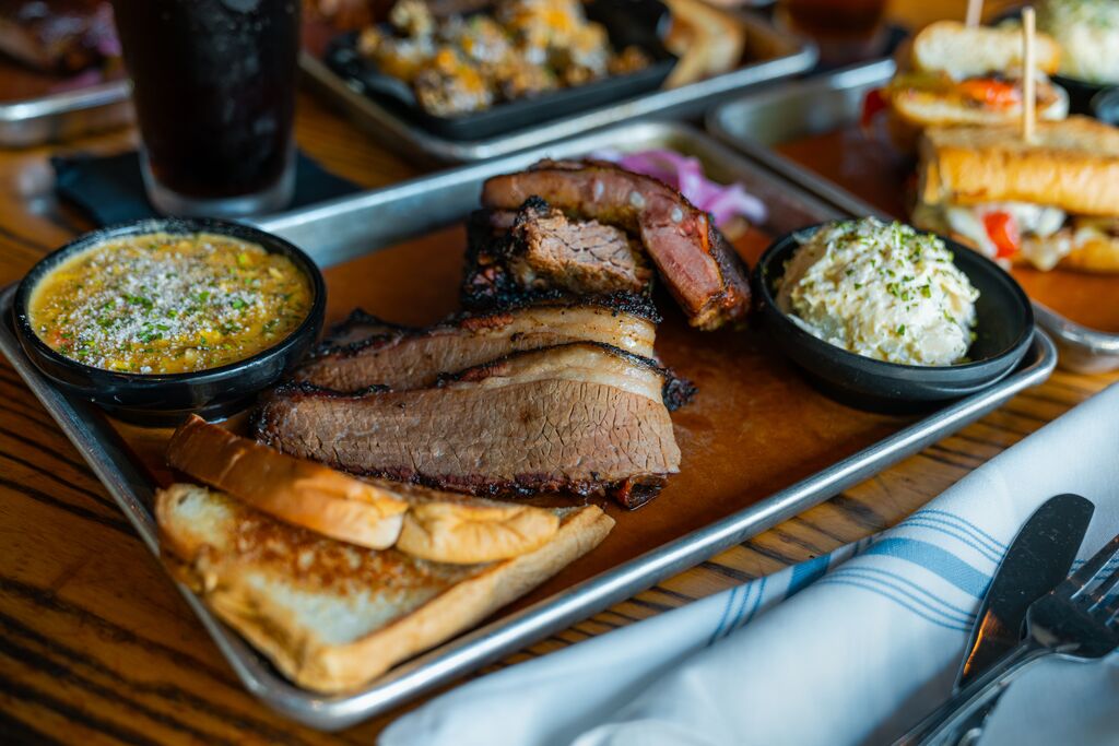 Several metal trays sit on a wood table with a variety of smoked meats and sides.